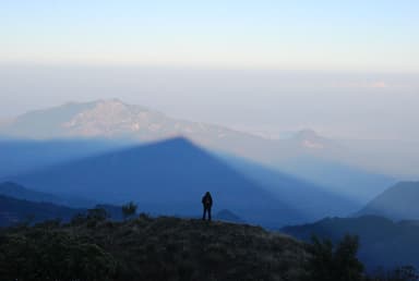 The shadow of Mount Ramelau cast over the hills at sunrise, with a hiker on the summit ridge