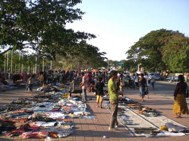 Goods laid out on the pavement at a busy street market under shade trees in Dili